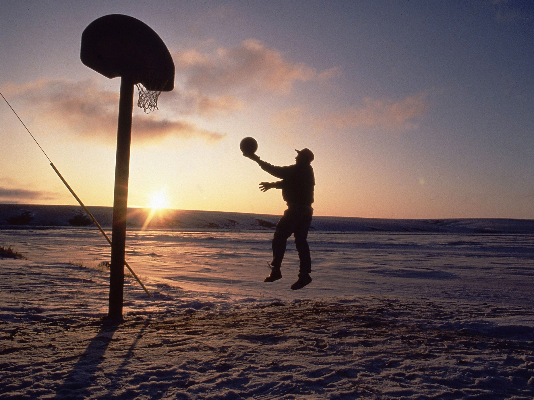 A lone basketball player shoots  as the sun peers over a ridge in Deering, Alaska.